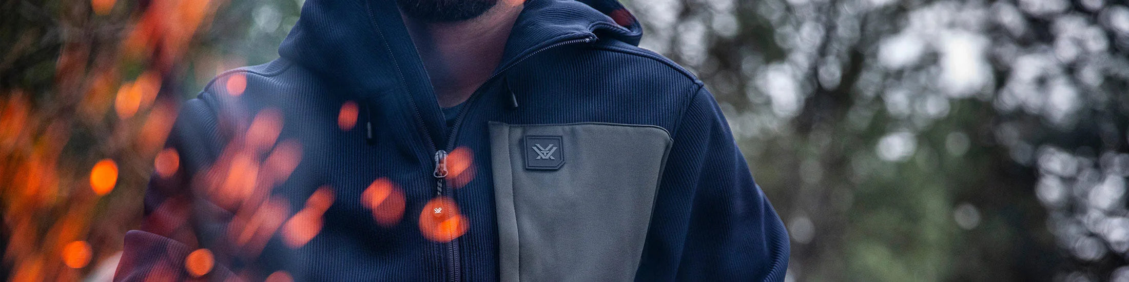Man wearing navy outdoor jacket by campfire with embers, forest background, apparel collection