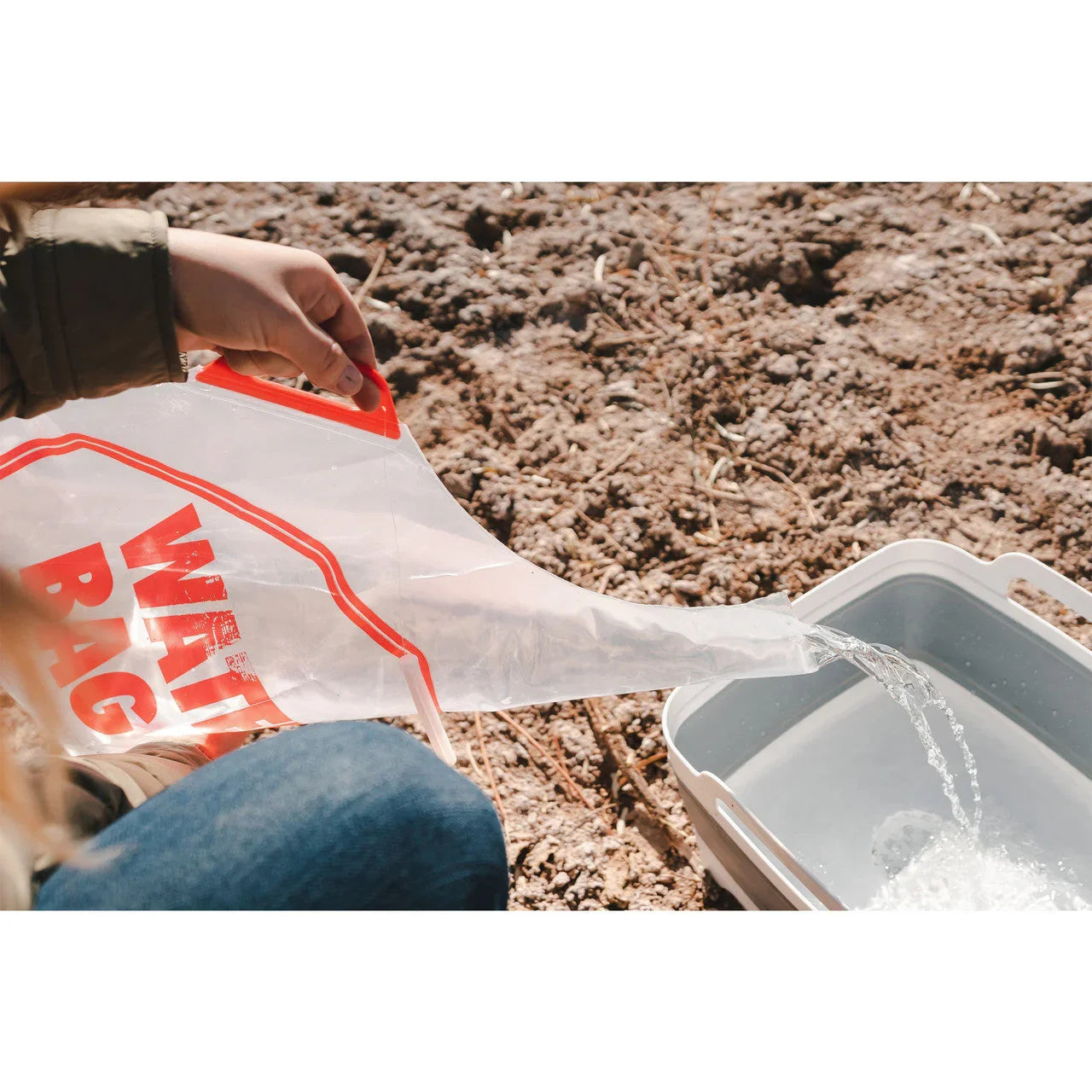 Person pouring water from clear water bag into a plastic container outdoors on dry soil