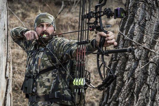 Camouflage archer aiming compound bow outdoors beside tree, bowhunting gear visible.