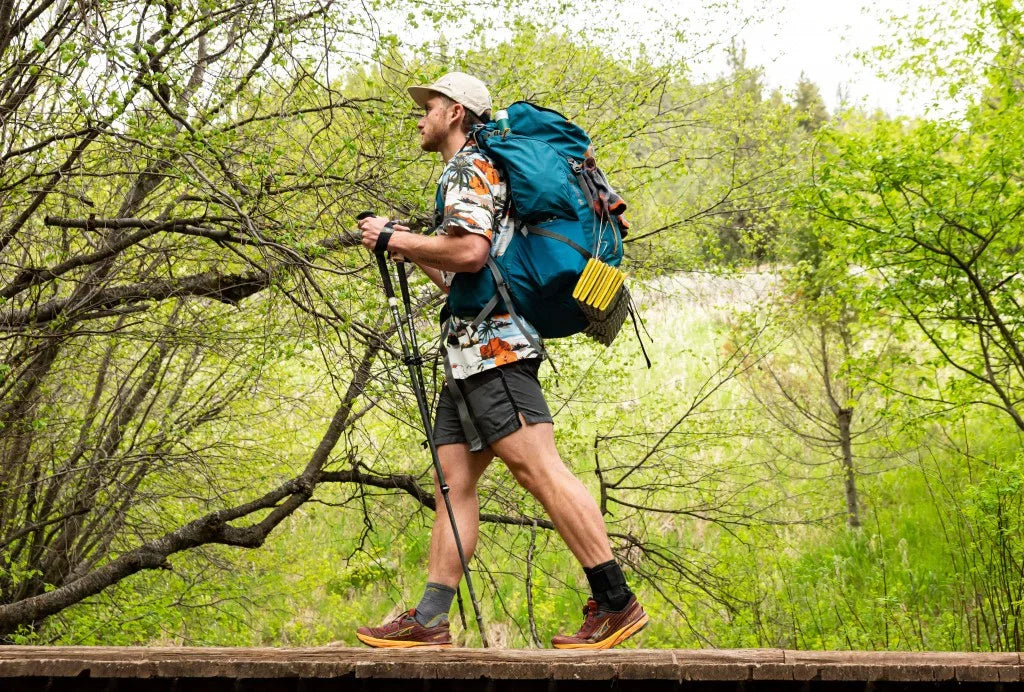 Man hiking in forest with large backpack and trekking poles, wearing outdoor gear and hat