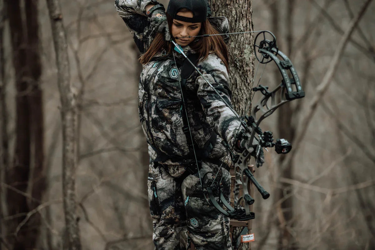 Woman in camo gear drawing a compound bow in a forest, archery hunting outdoors