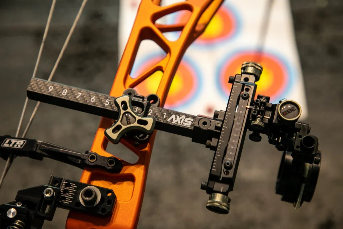 Close-up of compound bow sight with orange riser and blurred archery target in background