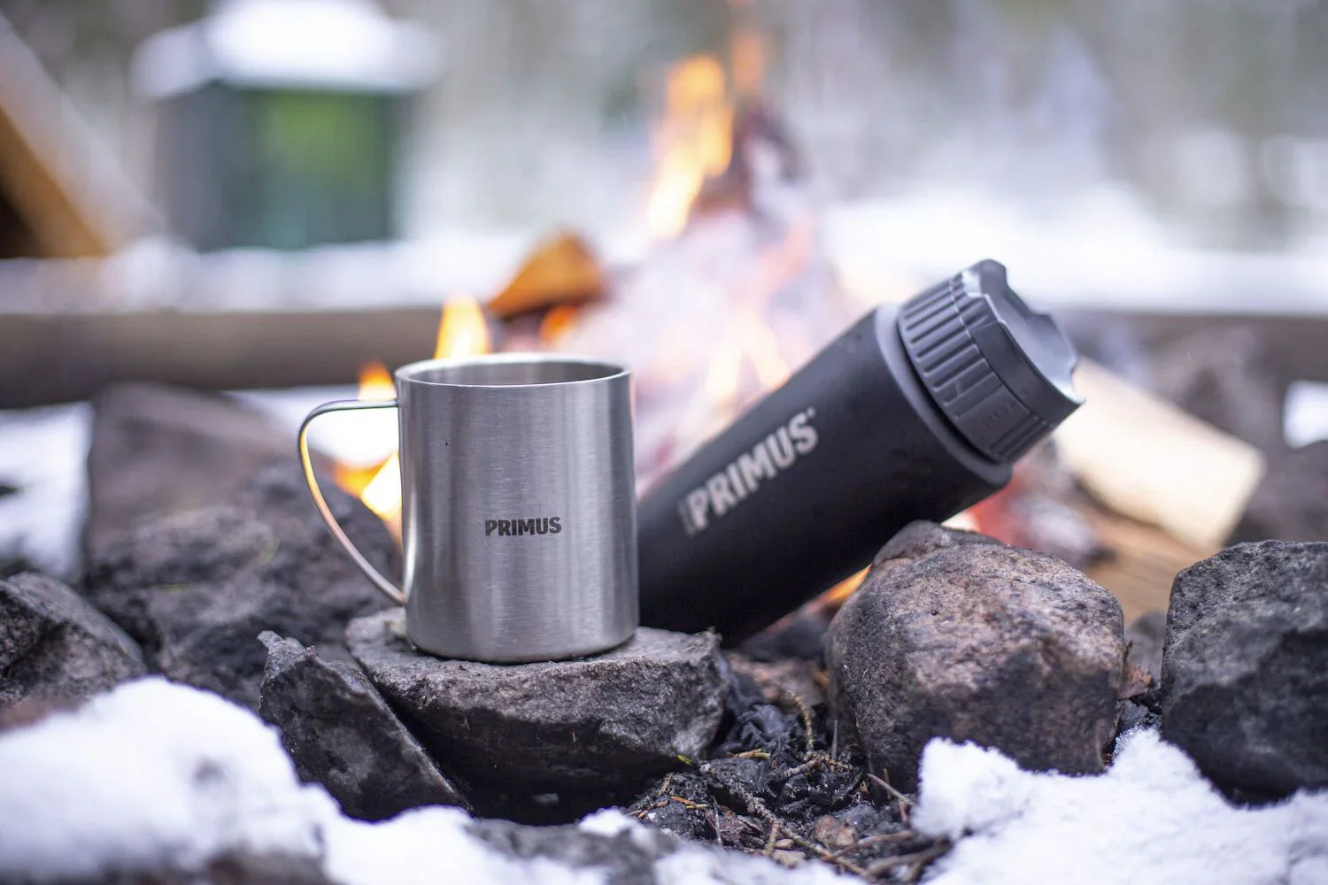 Primus metal mug and black thermos on rocks by snowy campfire outdoors