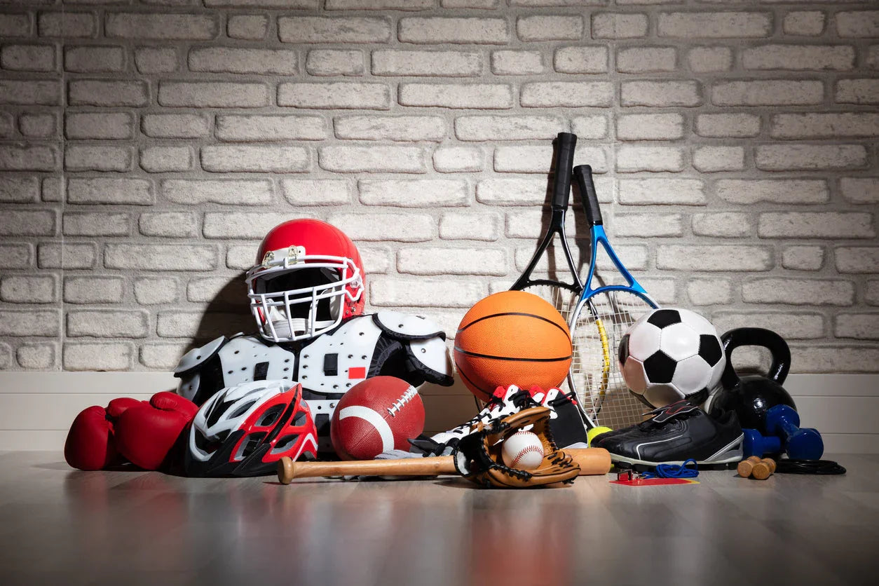 Assorted sports equipment including football helmet, basketball, tennis racquets, soccer ball and boxing gloves on wooden floor against brick wall.