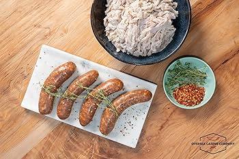 Raw sausage links on a marble tray with fresh herbs, sausage casings, and chili flakes on a wooden table.