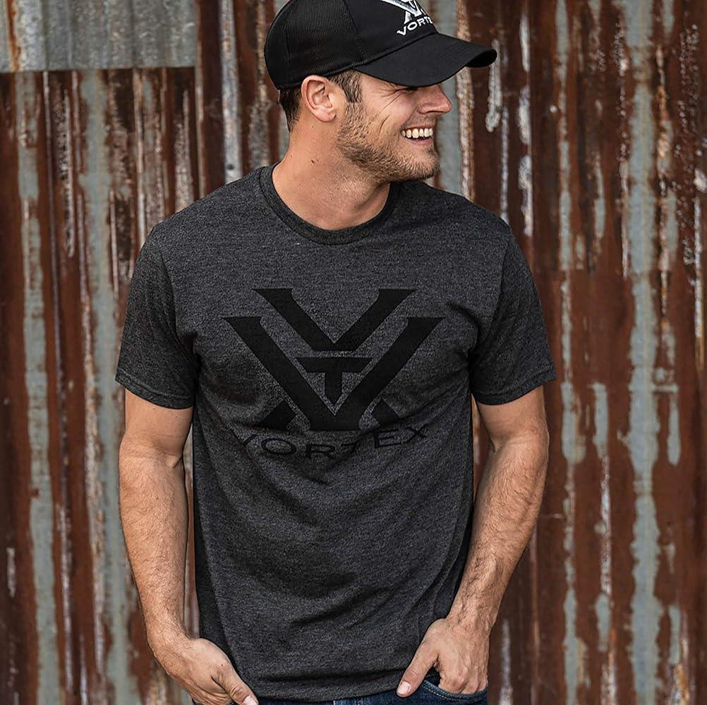 Man in Vortex logo t-shirt and cap standing by rusted metal wall, casual outdoor look