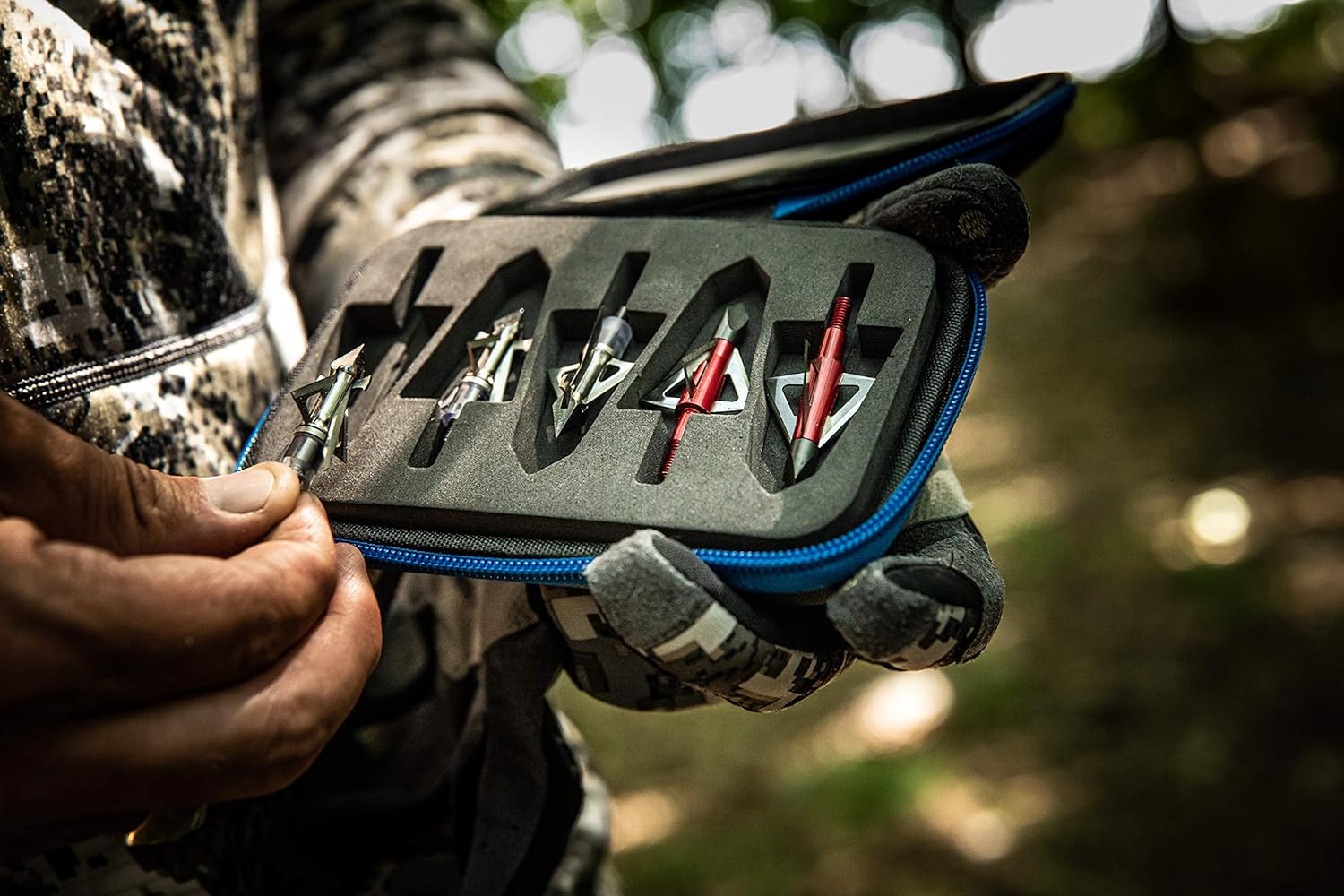 Hunter holding broadhead arrow tips in foam case with camouflage gloves outdoors