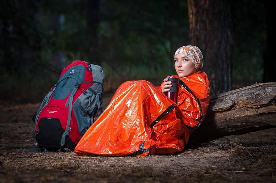 Woman in orange emergency sleeping bag holding mug, with hiking backpack in forest