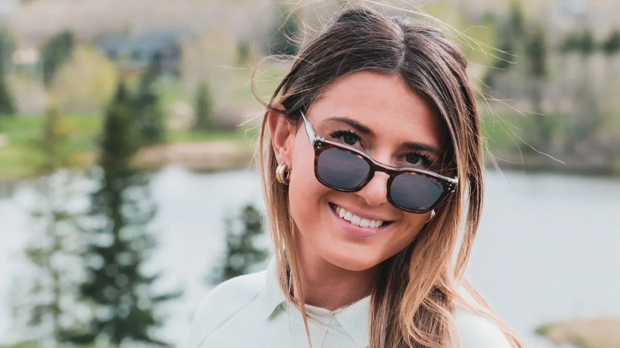 Smiling woman wearing sunglasses outdoors with trees and a lake in the background