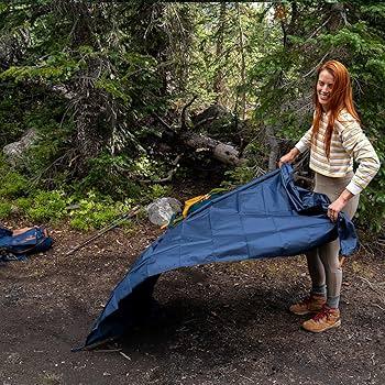 Woman setting up blue camping tarp in forest campsite, outdoor gear visible