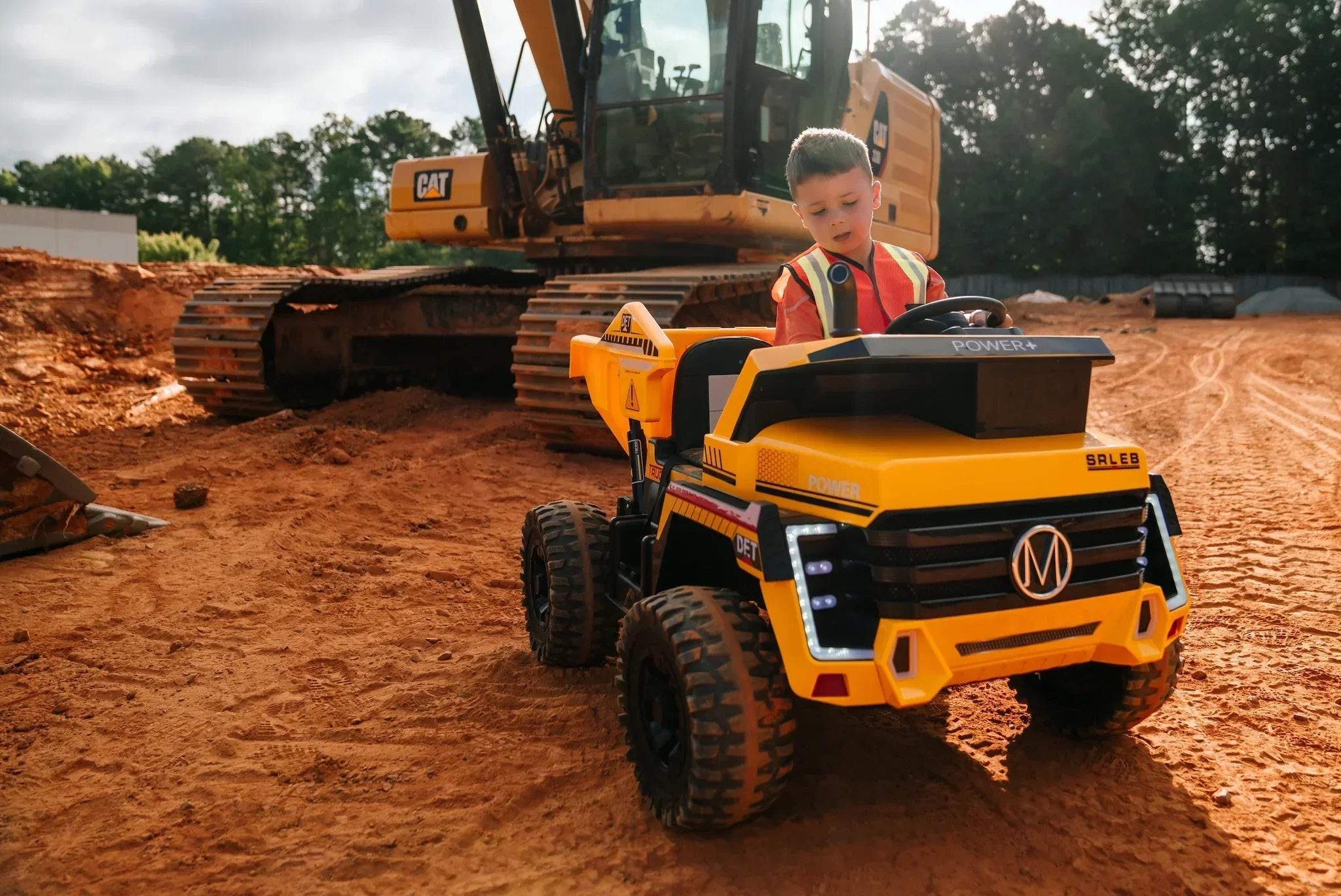 Child in safety vest driving yellow toy dump truck on construction site with excavator
