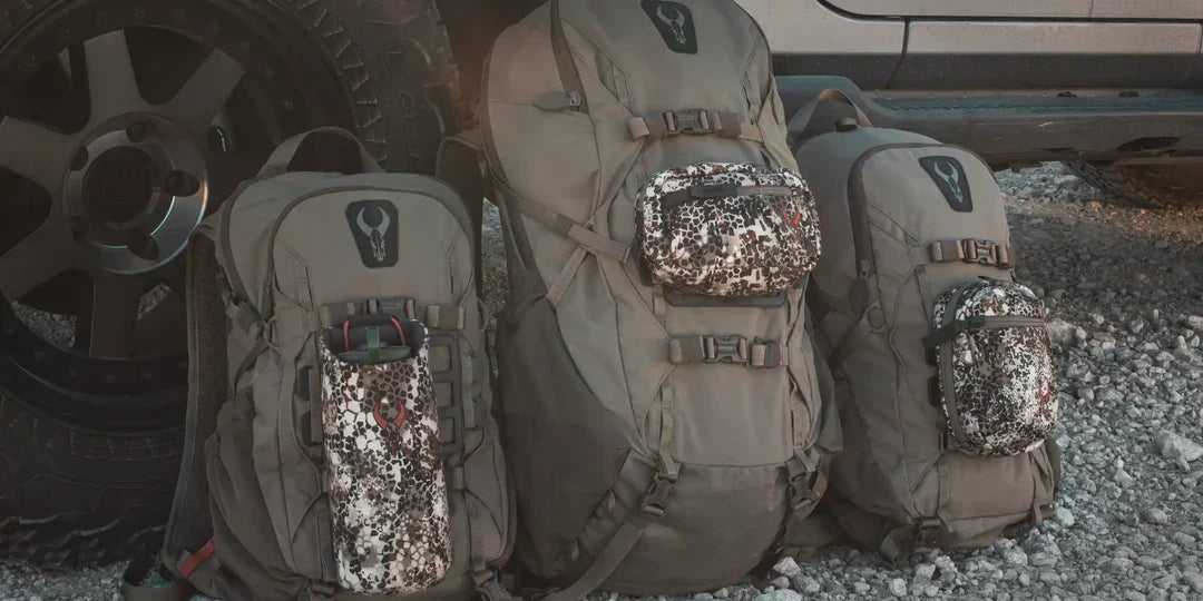 Three gray tactical backpacks with camo pouches on gravel beside a rugged vehicle tire