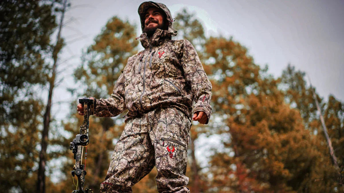 Man in camouflage hunting jacket and pants holding compound bow outdoors in forest
