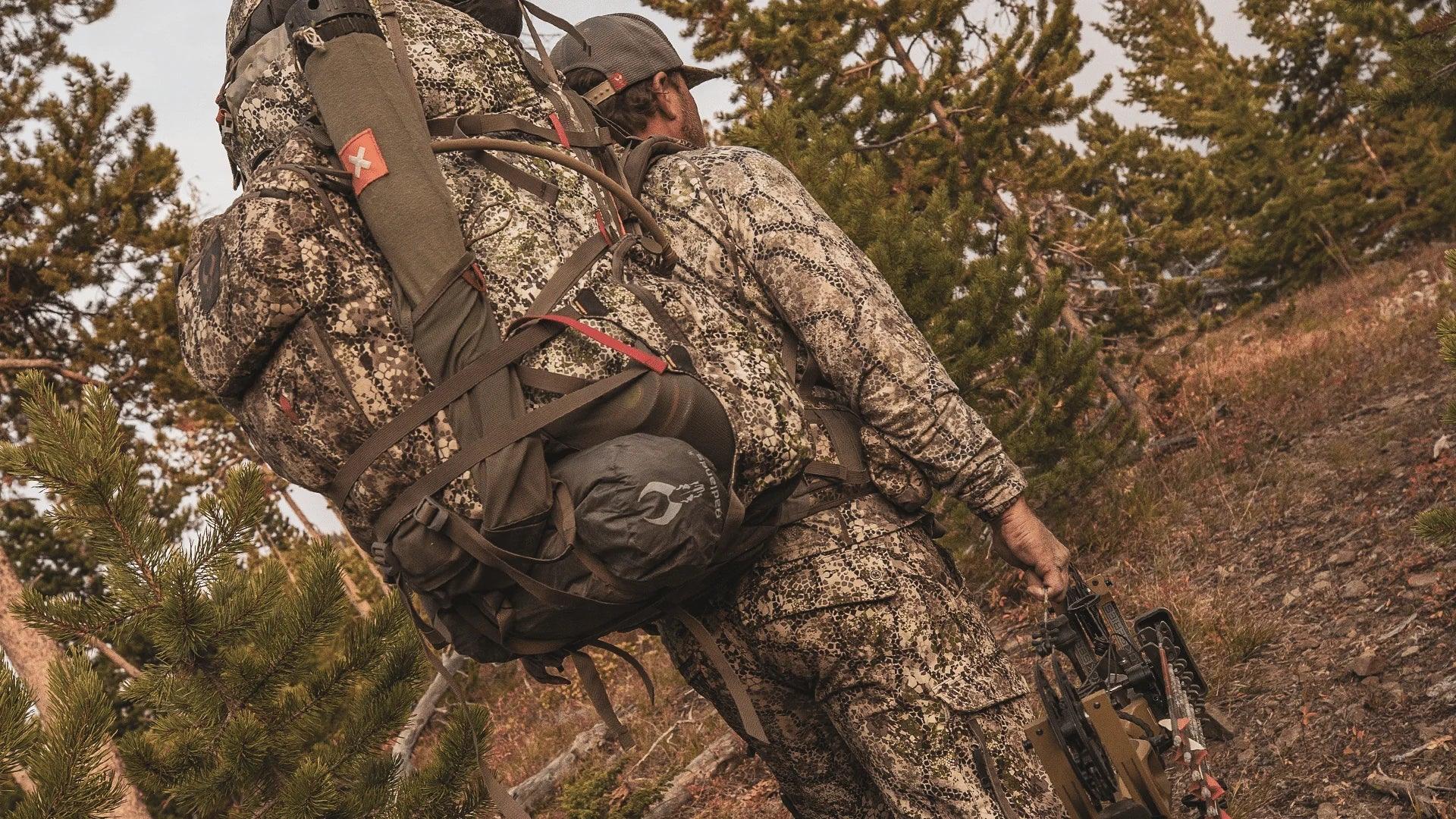 Hunter in camo gear with large backpack walking through pine forest outdoors