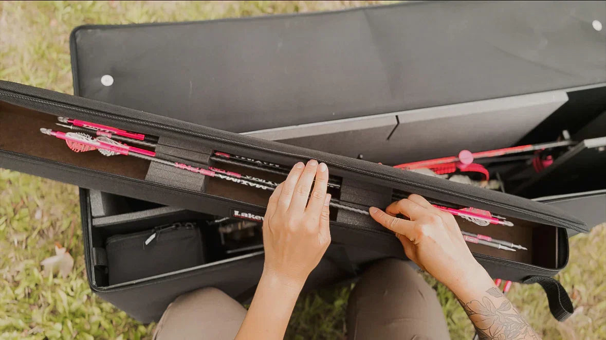 Person organizing pink and white arrows in an archery gear case outdoors on grass