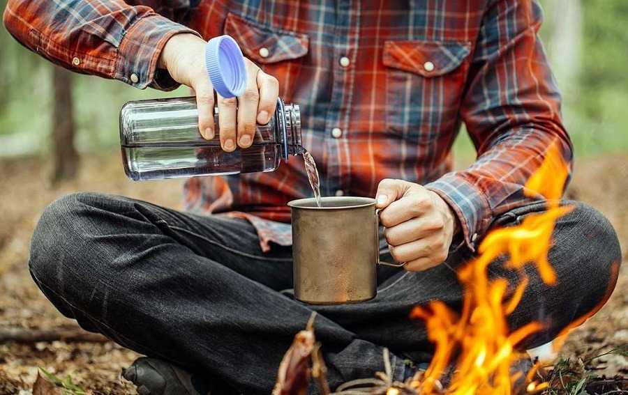Camper in plaid shirt pours water into mug by campfire in forest setting