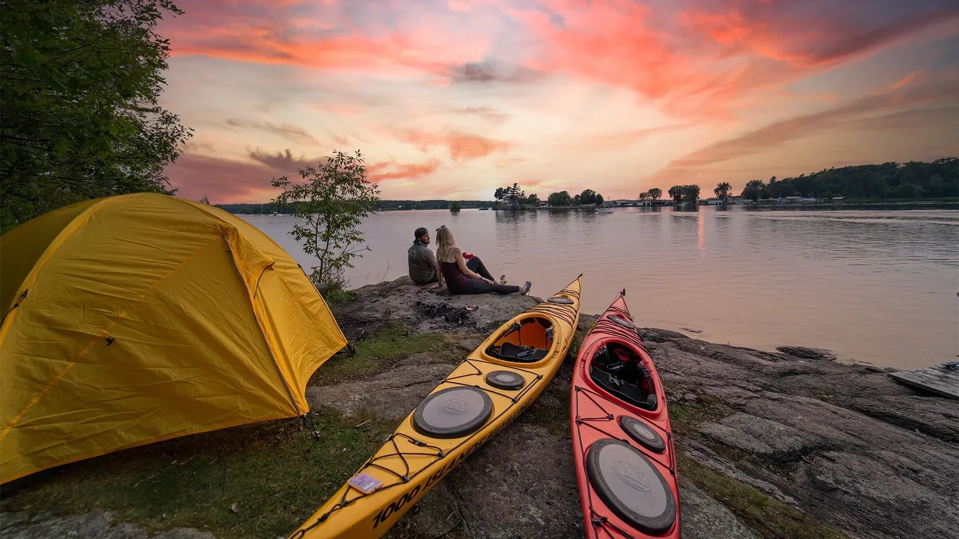 Yellow camping tent and kayaks by lakeside at sunset, couple sitting on rocks