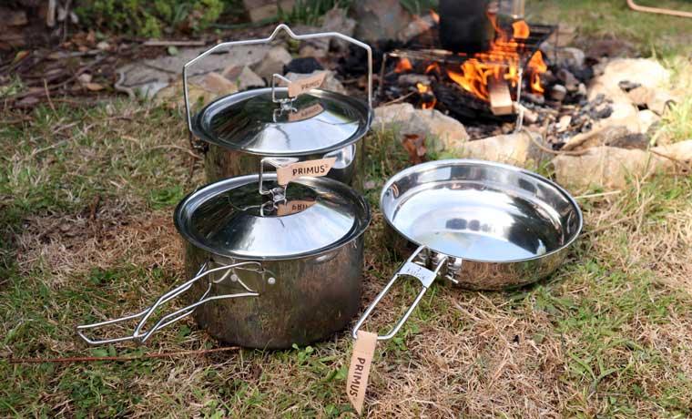 Stainless steel camping cookset with pots and pan near campfire on grass outdoors
