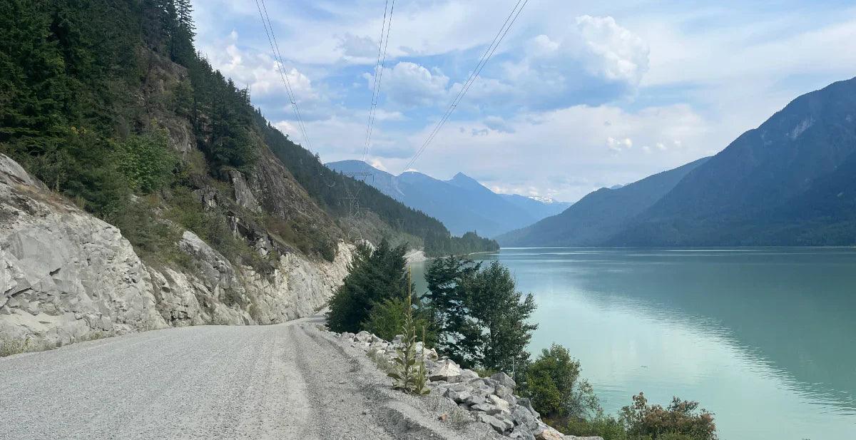 Gravel road beside mountain lake with forested cliffs and scenic blue sky landscape.