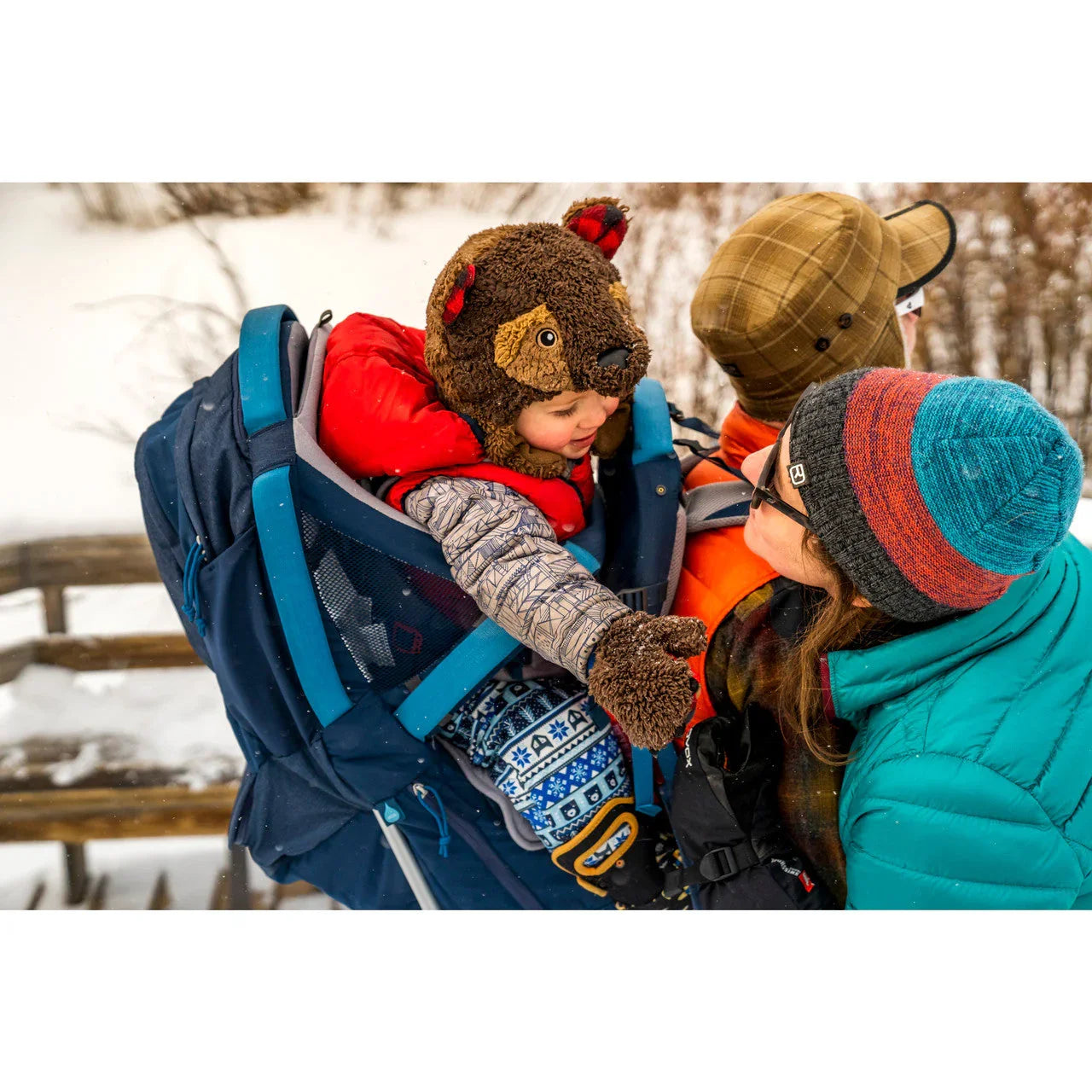 Child in bear hat riding in blue hiking carrier, winter family outdoor adventure, snow background