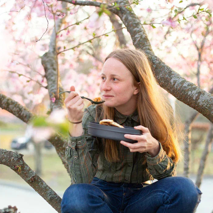 Woman eating from a black meal set while sitting in a blossoming cherry tree outdoors