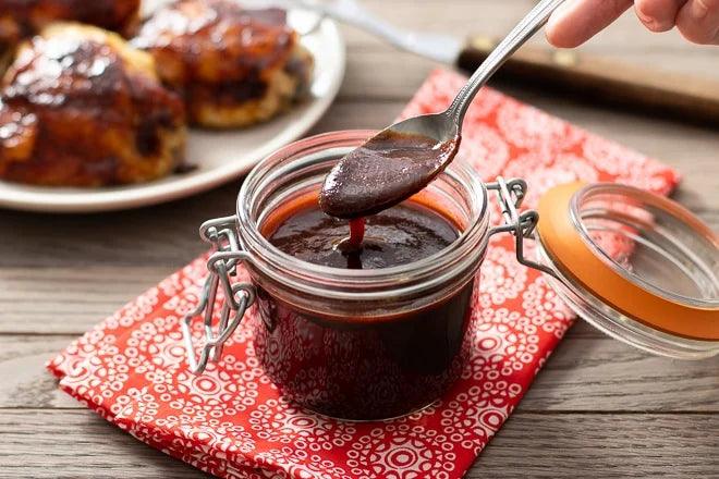 Jar of sweet smoky BBQ sauce on red cloth, spoon dipping, grilled chicken in background