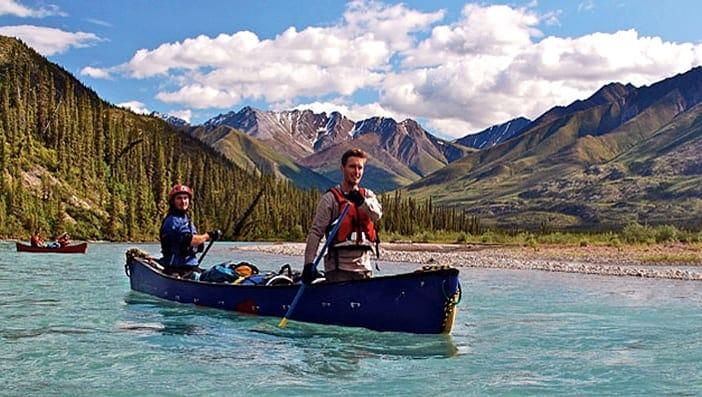 Two people canoeing on a clear Yukon River with mountains and pine trees in the background