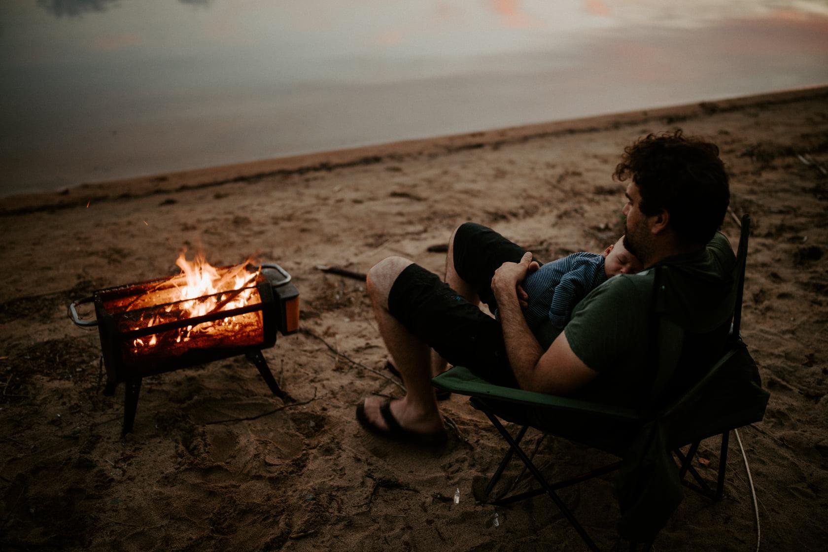 Man with baby relaxing in camping chair by Biolite portable fire pit on a sandy beach at dusk