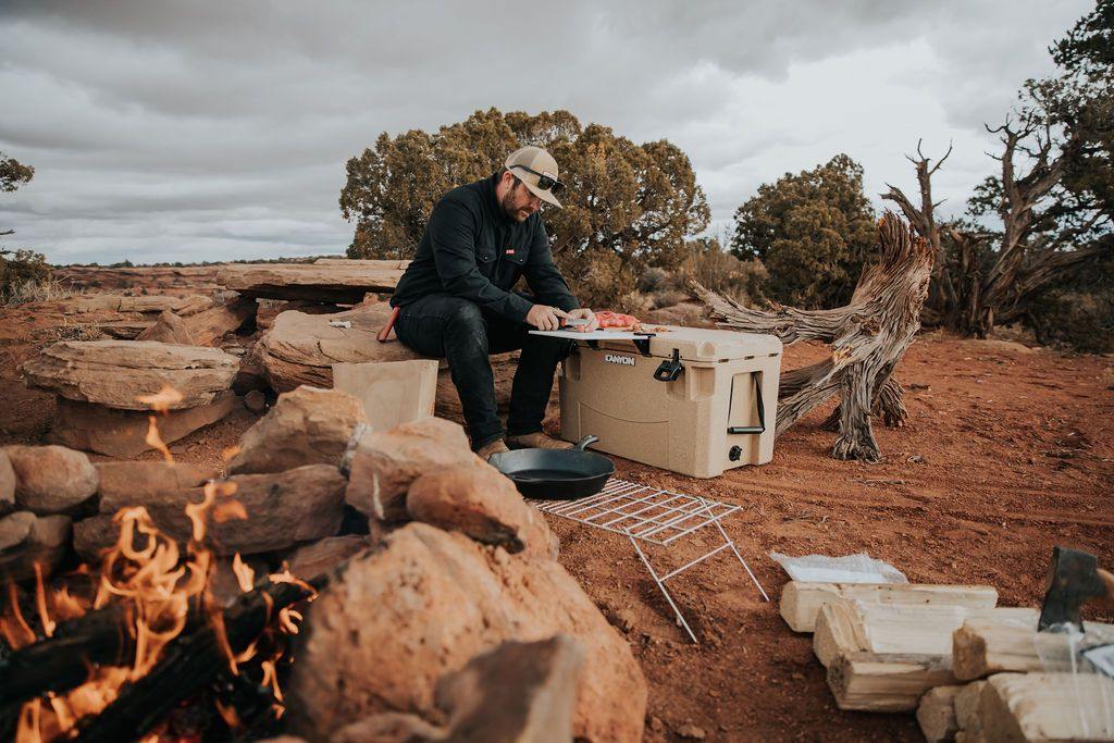 Man preparing food on Canyon Cooler at outdoor campsite with campfire and rugged desert landscape