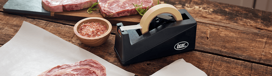 Butcher tape dispenser with meat, parchment paper, and bowl of salt on wooden table
