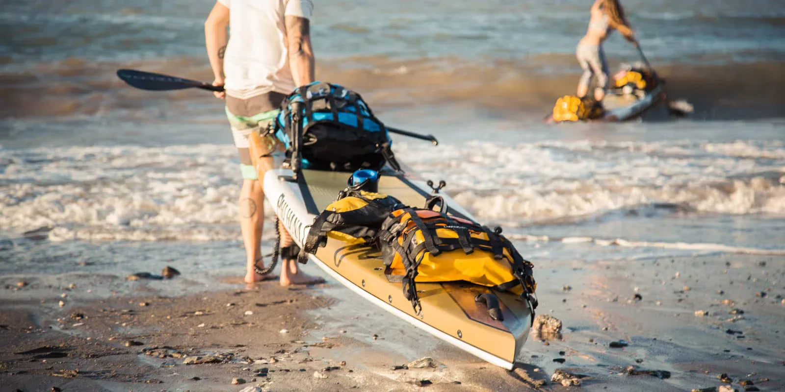 Paddle boarders with backpacks and dry bags on SUP boards at a sandy beach shoreline