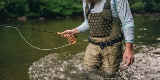 Man fly fishing in river wearing fishing waders and vest, surrounded by water and trees.