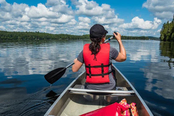 Woman kayaking on calm lake with red life jacket, surrounded by trees and blue sky