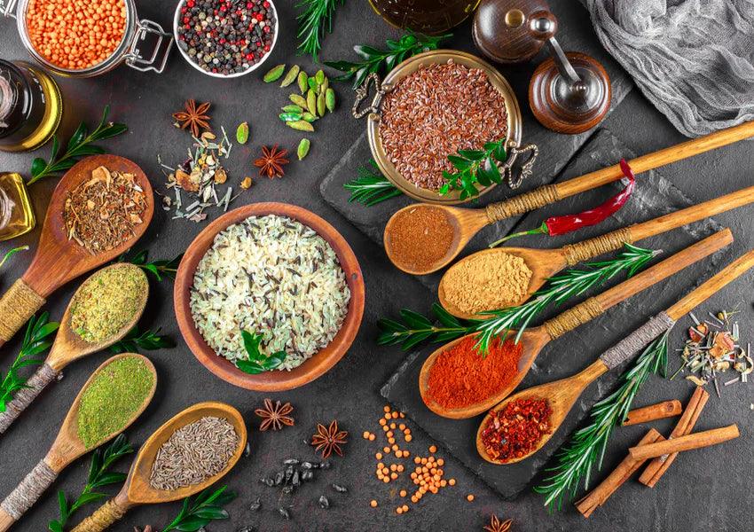 Assorted spices and herbs with rice in wooden spoons and bowls on dark kitchen table.