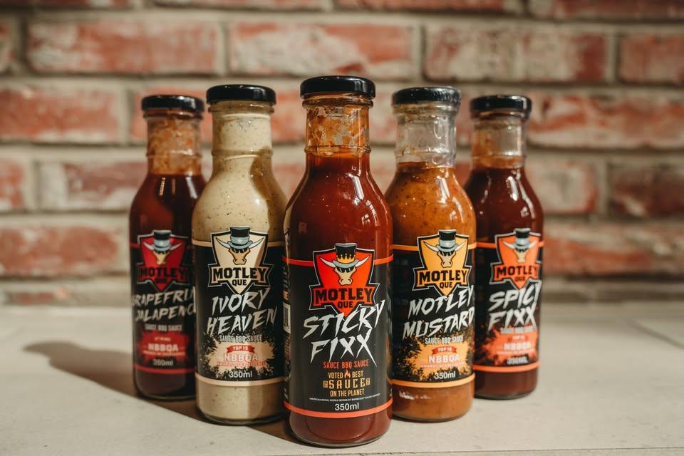 Motley Que BBQ sauces in bottles with various flavors on a kitchen counter, brick wall background