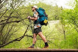 Man hiking with large Kelty backpack and trekking poles in green outdoor forest setting.