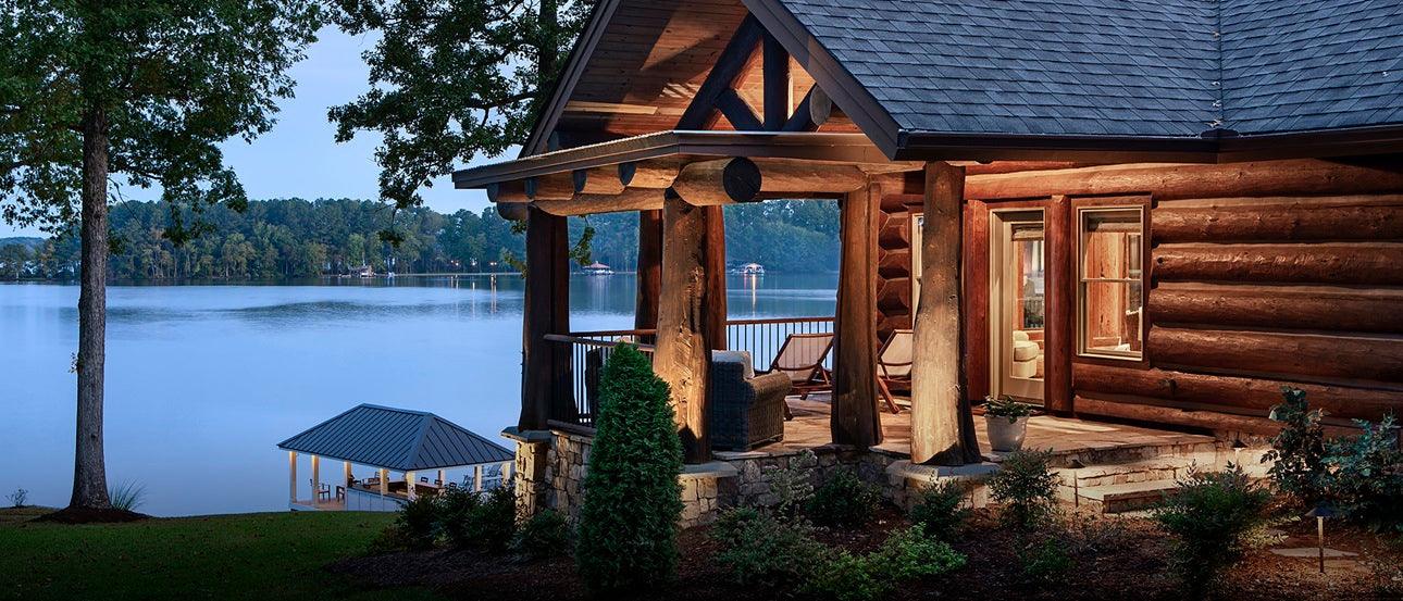 Rustic log home with covered porch overlooking a serene lake and dock at sunset