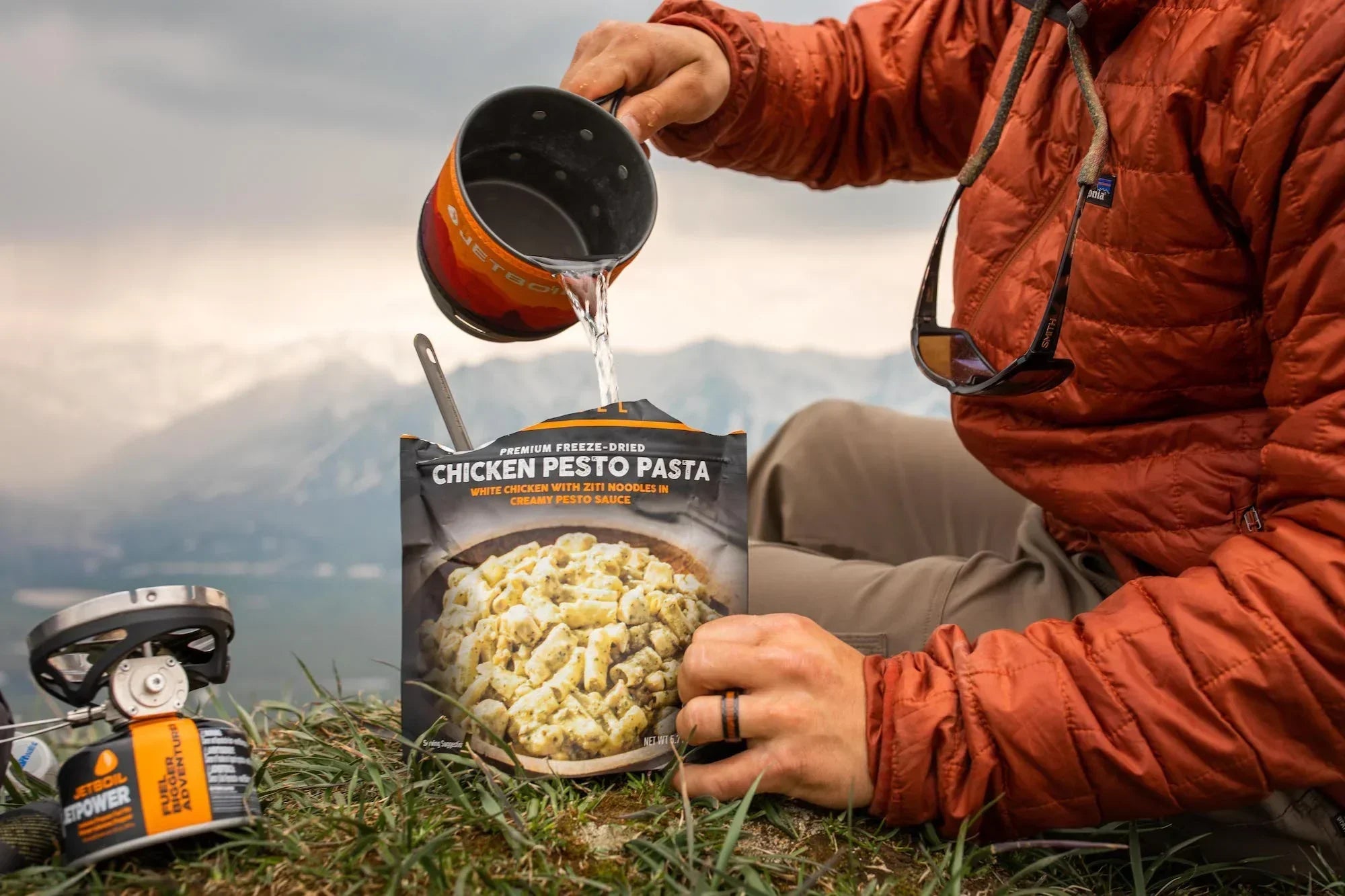 Camper pours water into Peak Refuel Chicken Pesto Pasta meal outdoors with mountains in background