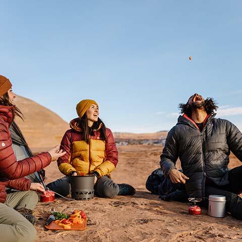 Three people in outdoor jackets camping on rocky terrain, cooking and eating with portable gear.