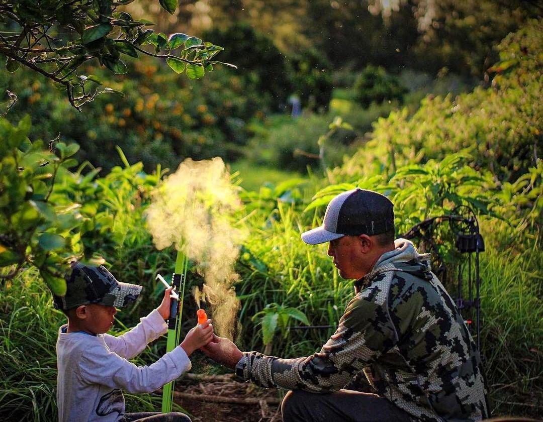 Man and boy using scent prevention spray outdoors, surrounded by green foliage, wearing camo hats and clothes.
