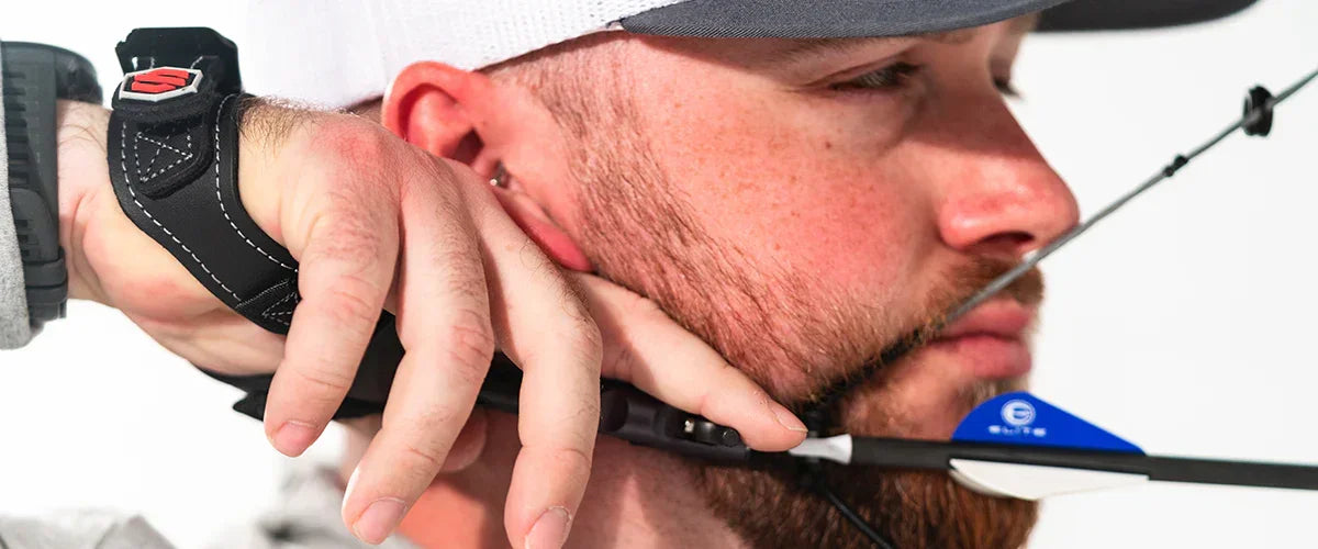 Man with beard aiming compound bow and arrow, wearing archery wrist release and cap