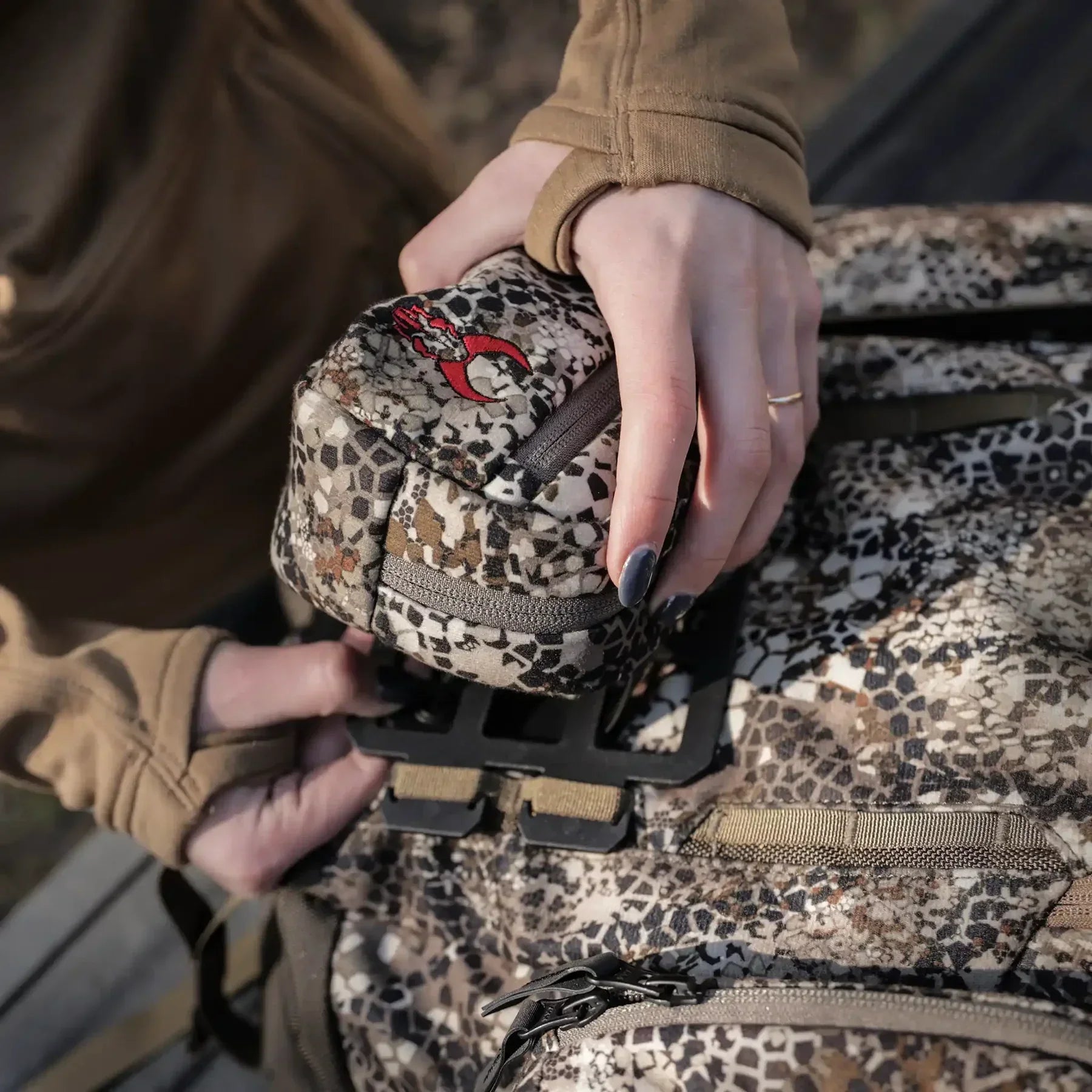 Hands attaching camo-patterned pouch to camouflage hunting backpack outdoors