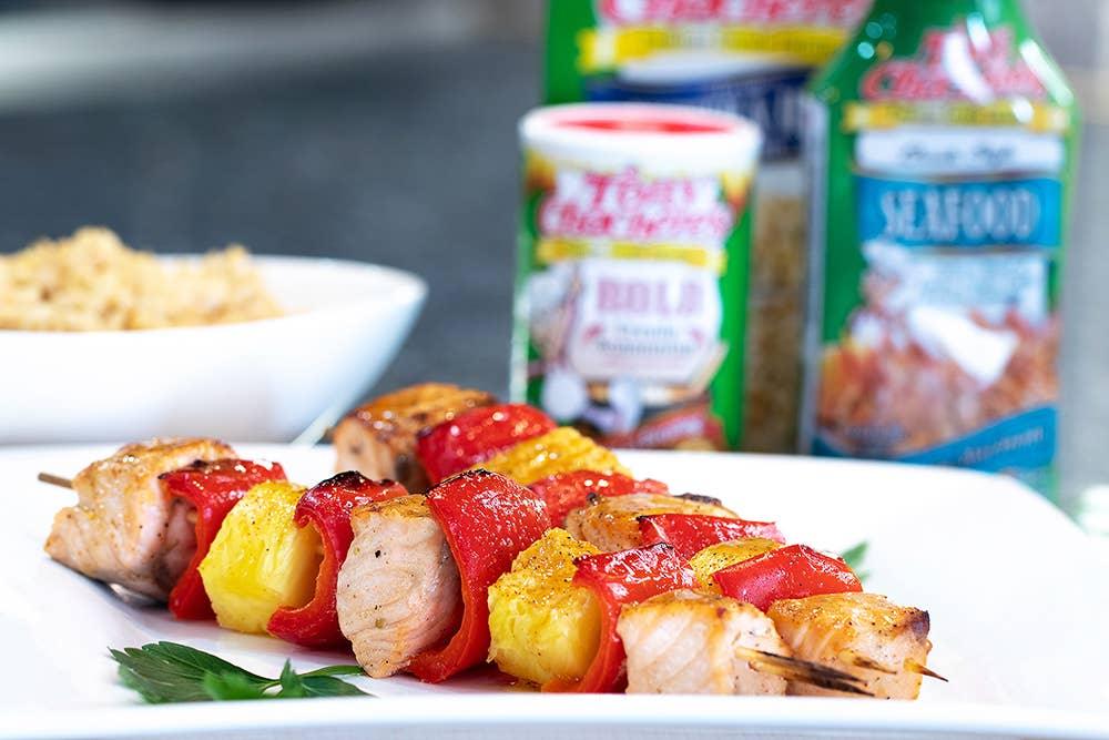 Grilled salmon, pineapple, and red pepper skewers on a white plate with seasoning bottles in background