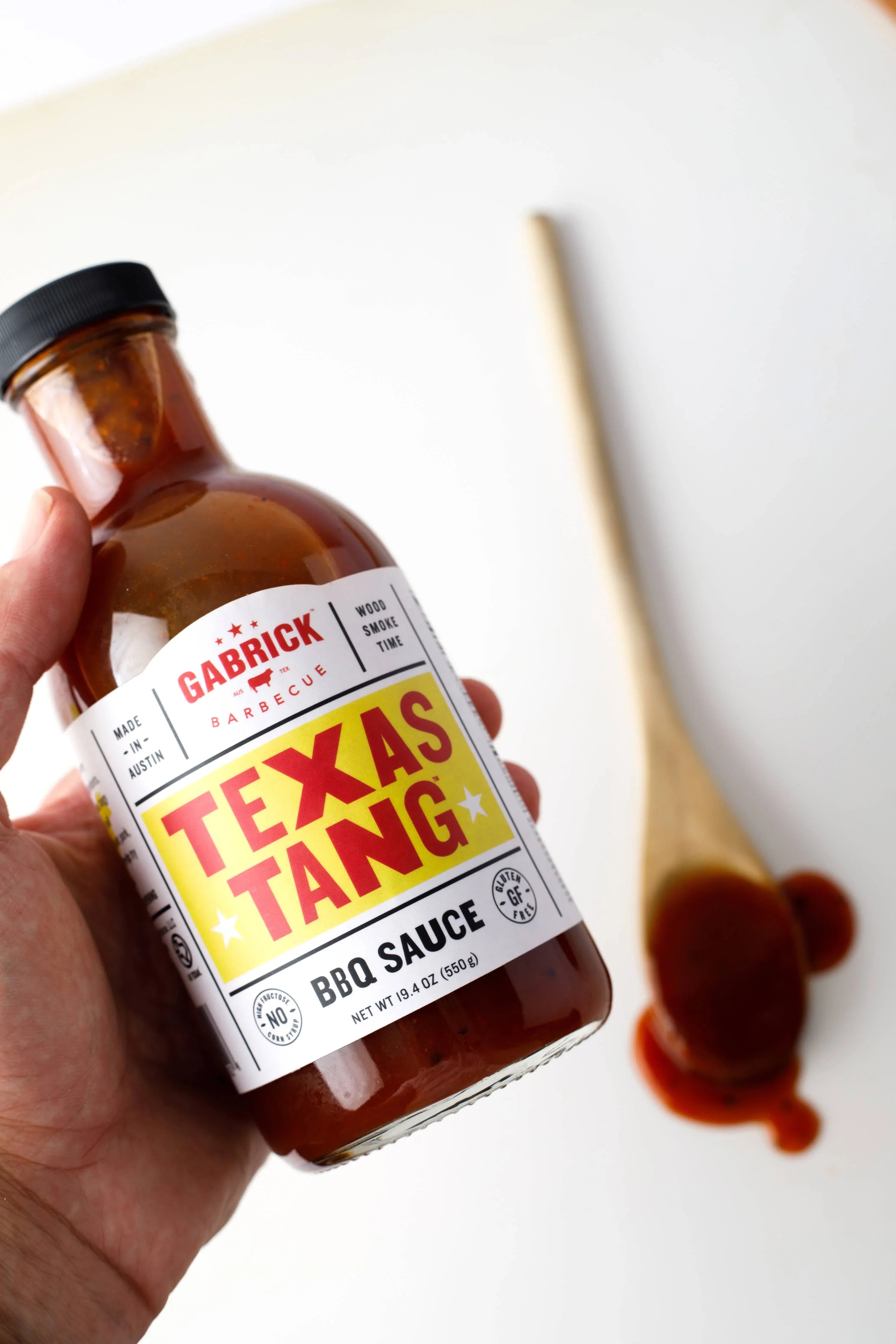 Hand holding Gabrick Texas Tang BBQ sauce bottle, wooden spoon with sauce on white background
