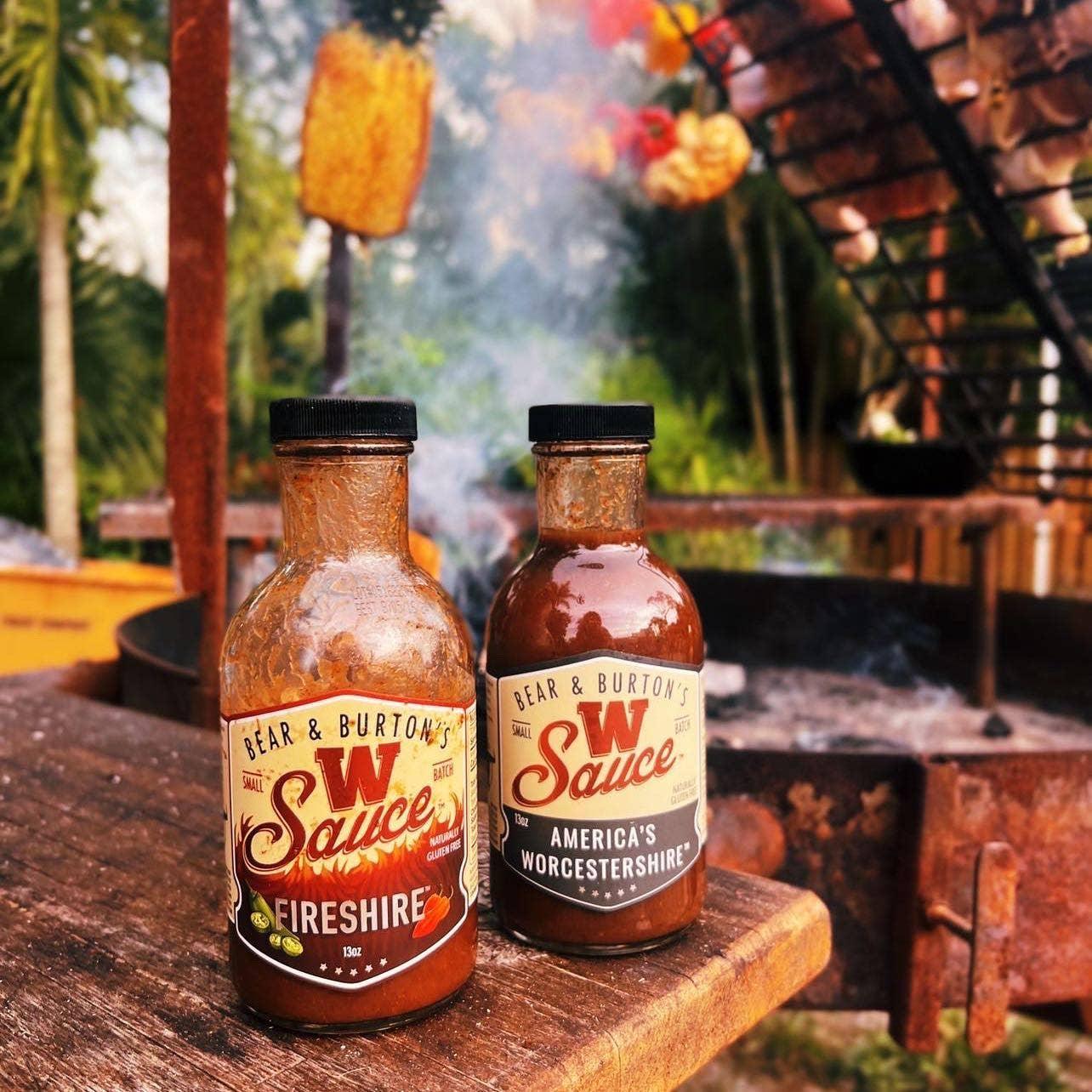 Bear & Burton's Worcestershire and Fireshire sauce bottles on outdoor wooden table near barbecue fire pit, grilling pineapple in background.