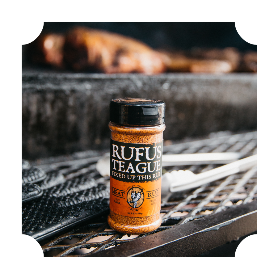 Rufus Teague meat rub seasoning bottle on a grill beside barbecue tools and cooked meat.