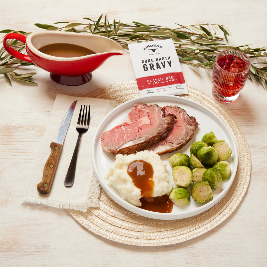 Plate of sliced prime rib, mashed potatoes with gravy, and Brussels sprouts, with bone broth gravy, a drink, and utensils on a light table.