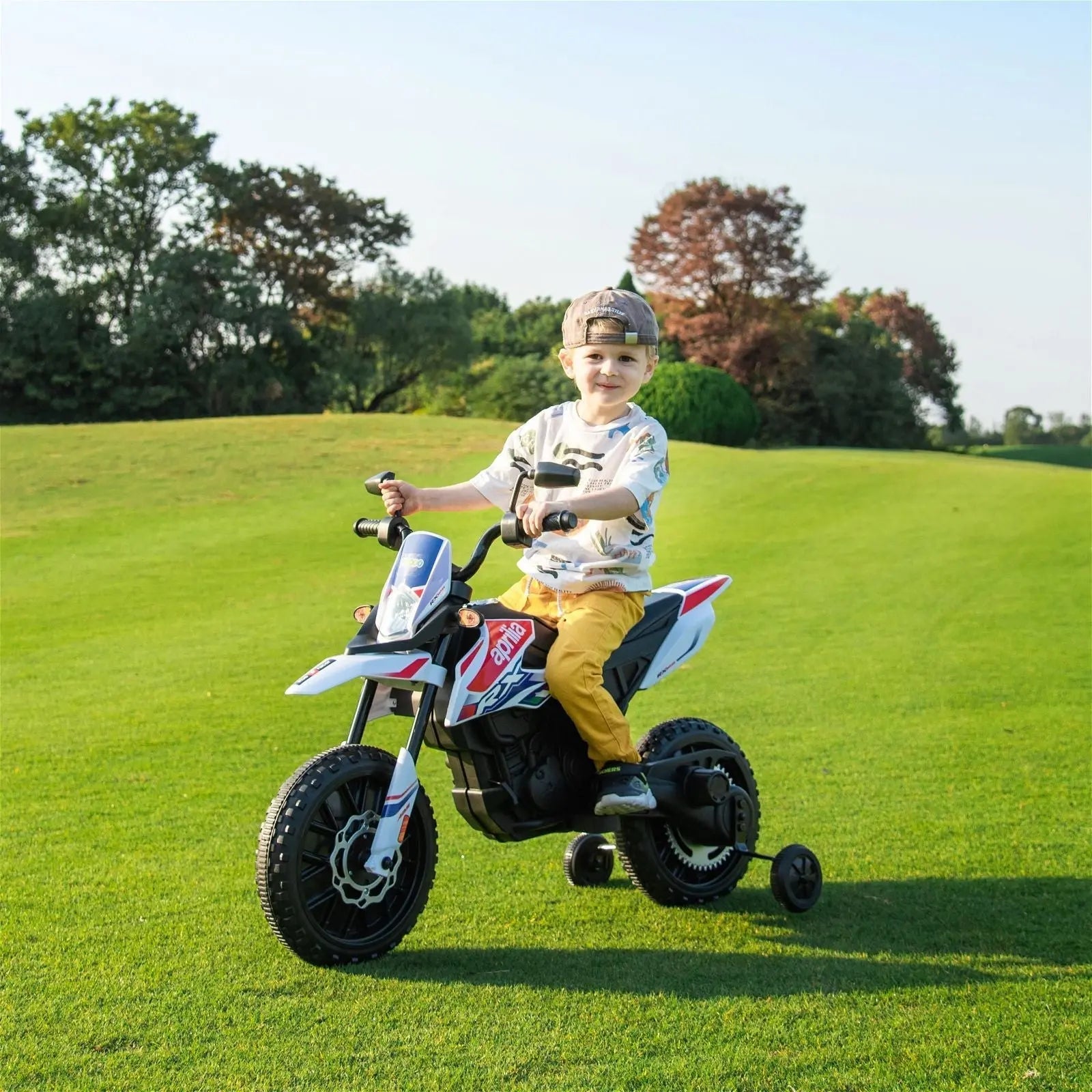 Child riding a 12V electric Aprilia motorcycle toy with training wheels on green grass