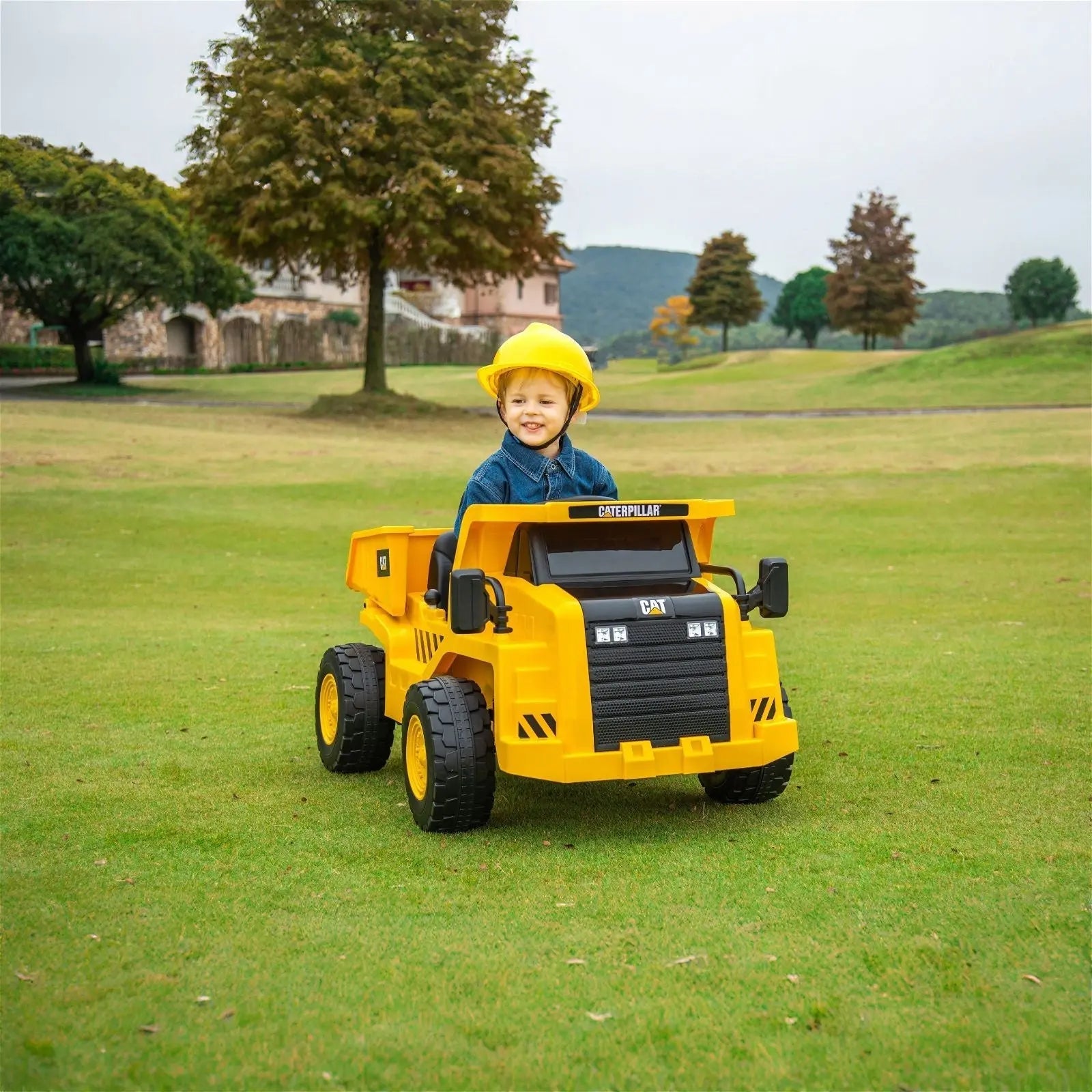 Child wearing a yellow helmet driving a yellow electric ride-on dump truck outdoors on grass.