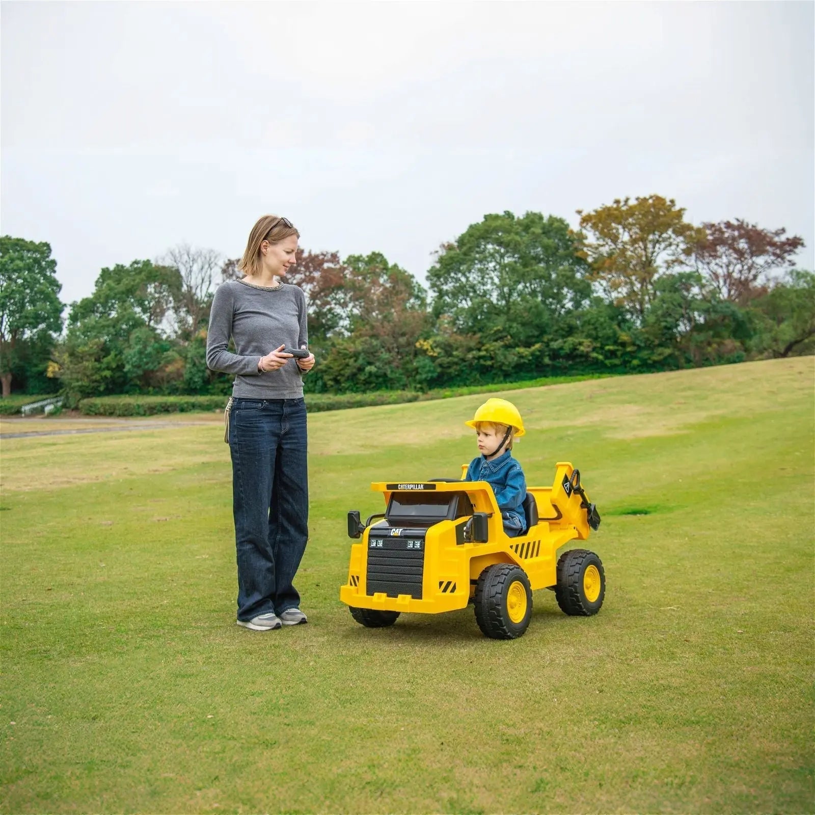 Child in yellow electric dump truck ride-on with hard hat, adult nearby, outdoors on grass